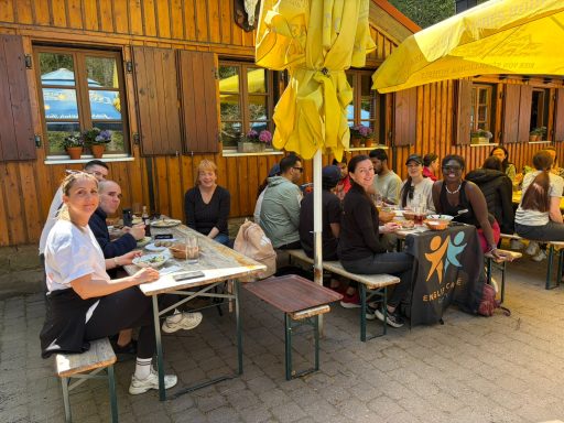 Gruppentreffen im Freien mit Personen an Tischen unter gelben Sonnenschirmen.