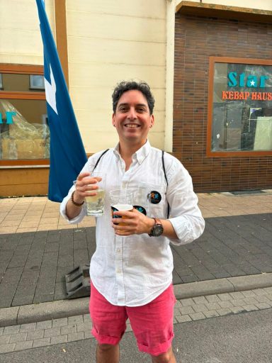 Junger Mann mit blauer Flagge und Getränk, bekleidet mit weißem Hemd und pinken Shorts.