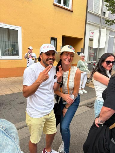 Zwei Personen posieren lächelnd für ein Foto auf einer Straße, umgeben von anderen Menschen.