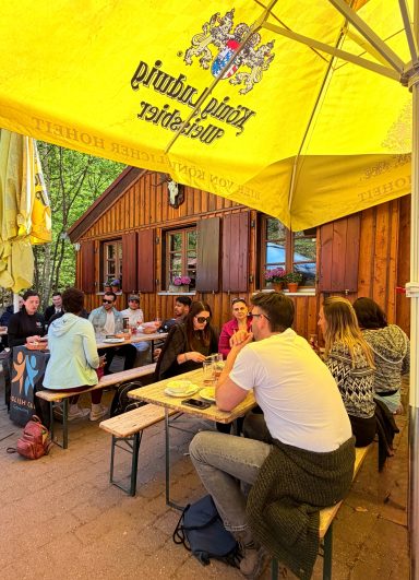 Gäste sitzen an Holztischen unter gelbem Sonnenschirm in einem Biergarten.