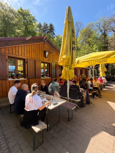 Menschen sitzen an Tischen vor einem Restaurant im Freien bei sonnigem Wetter.