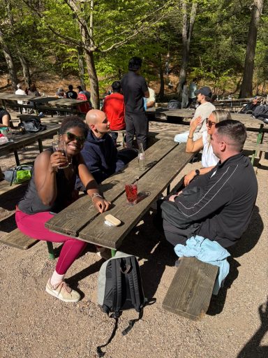 Gruppe von Personen sitzt an Tischen im Freien, umgeben von Bäumen und Natur.